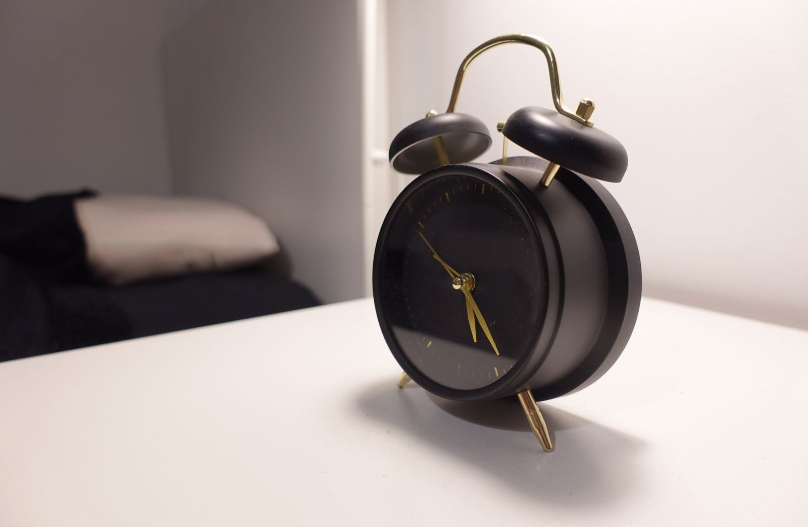 Black twin-bell alarm clock standing on a bedside table with a made bed behind it
